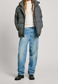 Giacca imbottita grigia con colletto alto, indossata sopra un maglione bianco a coste, abbinata a jeans larghi azzurri e stivaletti neri alla caviglia.