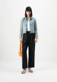 Ženska v svetlem jeans jakni, belem topu, črnih širokih hlačah, črnih sandalih in z oranžno torbico proti beli ozadju.