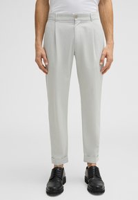 Pantaloni grigi chiari su misura con pieghe, slim fit e tasche frontali. Orli a cappio e fissati con un bottone. Abbinati a scarpe nere.
