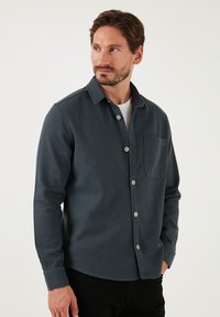 Camicetta in denim scuro con chiusura a bottoni sul davanti, taschino singolo sul petto e una texture sottile. Maniche lunghe con polsini abbottonati.