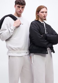 Νεαρός άνδρας με λευκό φούτερ που κρατάει μαύρο μπουφάν στον ώμο και νεαρή γυναίκα με σταυρωμένα τα χέρια που φοράει μαύρο μπουφάν bomber.