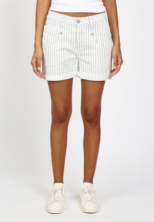 Frau trägt weiße, geraffte Shorts mit Nadelstreifen, weiße Turnschuhe und ein weißes, bauchfreies Oberteil, steht vor einem schlichten Hintergrund.