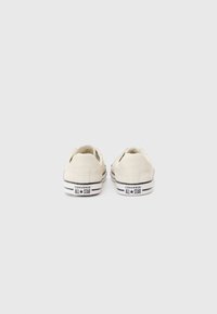 Beige canvas low-top sneakers with black rubber soles, featuring a logo at the heel and white laces. Simple design with minimal detailing.