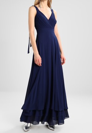 Kvinde iført en ærmeløs, mørkeblå V-hals kjole med plisseret krop og lagdelt nederdel, kombineret med sølvfarvede spidse hæle.