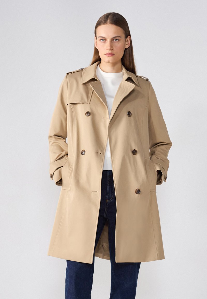 Beige trenchcoat, jossa on kaksirivinen malli, kaksi etupussia ja nappiyksityiskohtia; valmistettu sileästä kankaasta ja istuvuudeltaan muotitettu.