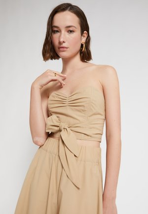 Giovane donna con capelli castani corti che indossa un top beige senza spalline con un fiocco e pantaloni a vita alta abbinati su uno sfondo semplice.