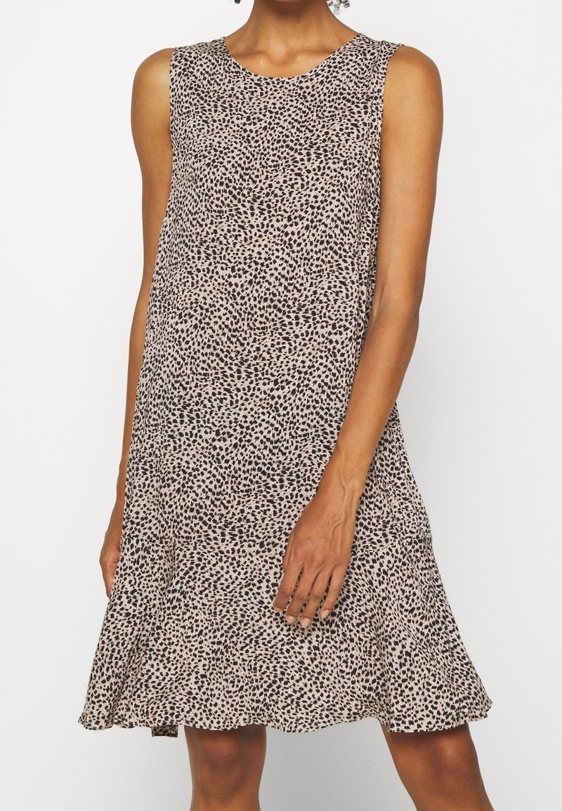 Robe beige sans manches avec des points noirs irréguliers, coupe ample, longueur au genou, portée par une personne debout sur un fond blanc uni.