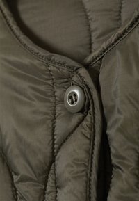 Close-up of dark green quilted fabric with stitching, featuring a large matte button sewn onto the material.