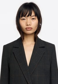Blazer de cuadros negros con un escote en V profundo, hombros estructurados y una tela texturizada con acentos marrones. Diseño minimalista, sin botones.