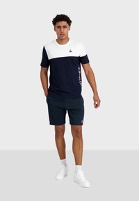 Camiseta de manga corta en azul marino y blanco con un diseño de bloques de color horizontales, que presenta un logotipo y un detalle de texto en el lateral, combinada con pantalones cortos azul marino.