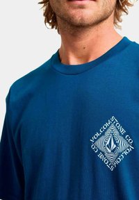 Hombre con una camiseta azul de Volcom con un logo geométrico y texto en blanco en el lado izquierdo del pecho, cortada por debajo de la nariz.