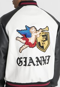 Læder varsityjakke med sorte ærmer og hvid krop. Ryggen har en farverig engel broderi og "GIANNI" i fed skrift.