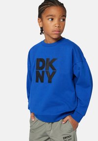 Blaues Sweatshirt mit einem schwarzen "DKNY"-Logo, Rundhalsausschnitt und lässiger Passform. Getragen mit hellgrauen Hosen.
