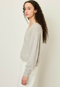 Pull en maille gris clair, oversize, avec un décolleté en V et des manches longues. Fabriqué en tissu doux et texturé. Associé à une jupe blanche avec une poche.