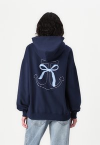 Mörkblå huvtröja med en stor vit och blå rutmönstrad ankare och rosett grafik på baksidan. Har en justerbar huva och ribbade ärmslut.