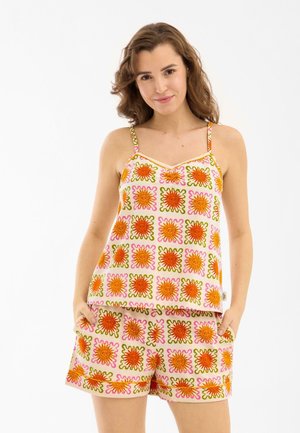 Young woman wearing sleeveless top and shorts with bright orange sun patterns, standing with hands in pockets, smiling gently.