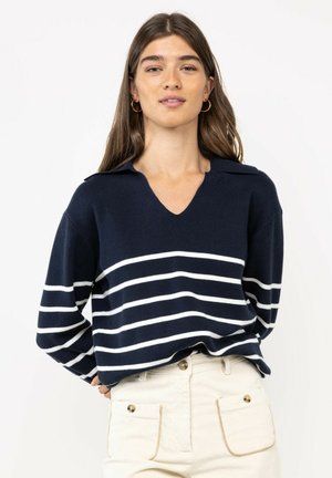 Joven mujer con cabello largo y castaño, vistiendo un suéter azul marino con rayas horizontales blancas y pantalones de tiro alto color crema con bolsillos.