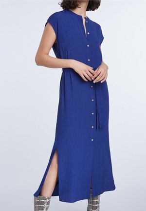 Vrouw draagt een blauwe mouwloze jurk met knopen, zijsplitten en zilverkleurige enkellaarsjes met slangenhuidmotief op een lichte achtergrond.