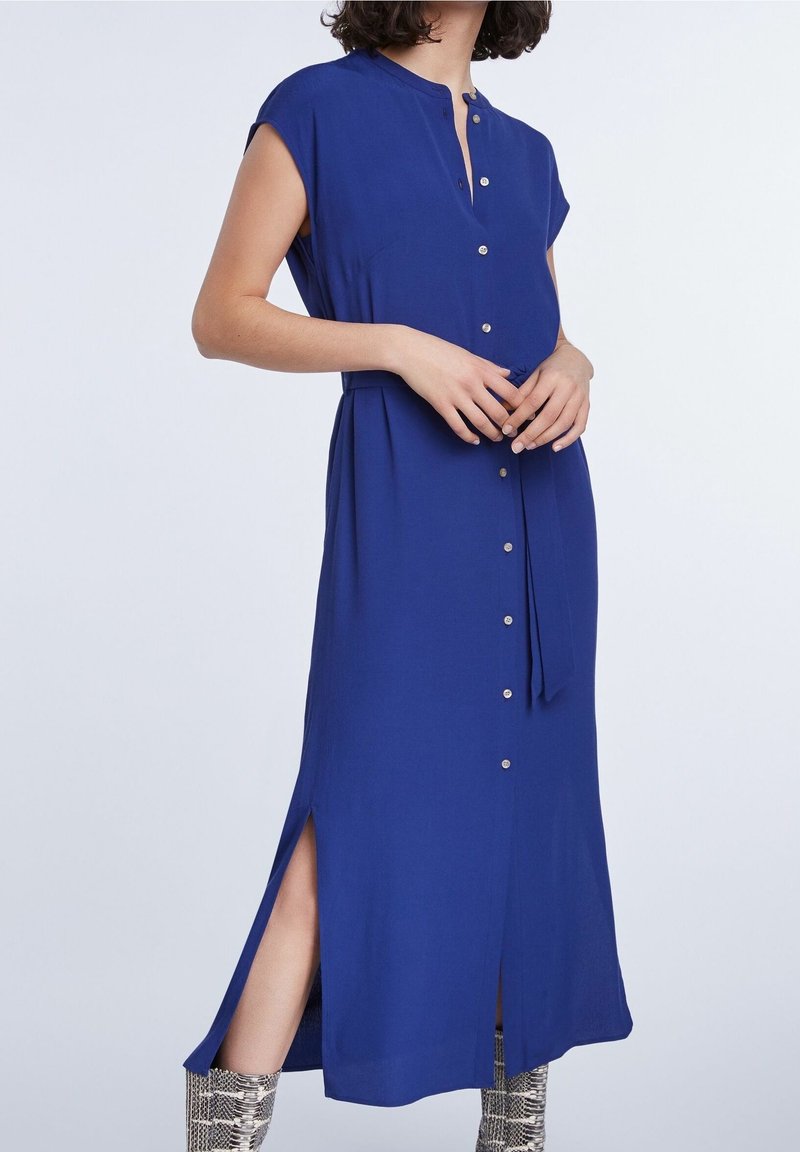 Femme portant une robe bleue sans manches boutonnée avec des fentes latérales et des bottines argentées à motif peau de serpent sur un fond clair.
