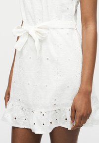 Λευκό φόρεμα με σχέδια eyelet με λουλουδένιες οπές, φιόγκο στη μέση και βολάν στο τελείωμα, φορεμένο από κάποιον με μεσαία σκούρα χροιά δέρματος.