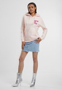 Rosa Kapuzenoberteil mit dem Text "WARTEN AUF DEN FREITAG"; Vorgentasche, kombiniert mit einem Denim-Minirock, Netzstrümpfen und silbernen Stiefeletten.