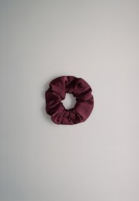 Donkerpaarse stoffen scrunchie met geplooide textuur geplaatst op een effen lichtgrijze achtergrond.