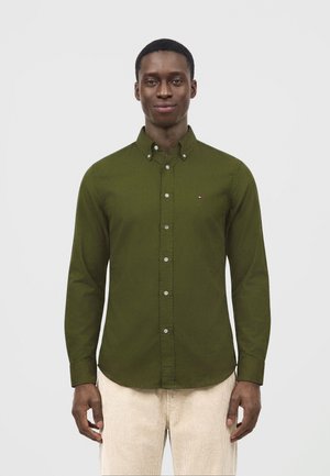 Homme portant une chemise boutonnée vert olive et un pantalon beige clair, debout devant un fond blanc uni.