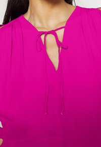 Femme portant un chemisier rose vif avec un détail à nouer au cou et une petite ouverture en forme de trou de serrure à l'encolure.
