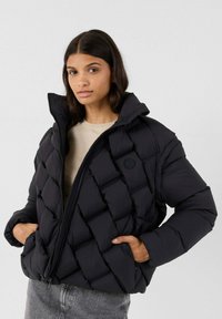 Jonge vrouw met donker haar draagt een zwarte gewatteerde pufferjack over een beige top, met haar handen in de jaszakken tegen een neutrale achtergrond.