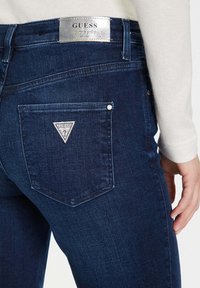 Jeans in denim blu scuro con un'etichetta argentata "GUESS" sulla cintura, caratterizzati da un logo a triangolo sulla tasca e dettagli di cuciture a contrasto.
