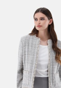 Hellgraue, gewebte Tweedjacke mit fransigen Kanten, offener Vorderseite und zwei aufgesetzten Taschen, getragen über einem weißen Oberteil.