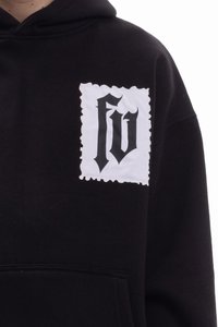 Sudadera con capucha negra con un parche blanco de borde irregular que muestra un logo en fuente estilizada negra. El material parece suave y texturizado.