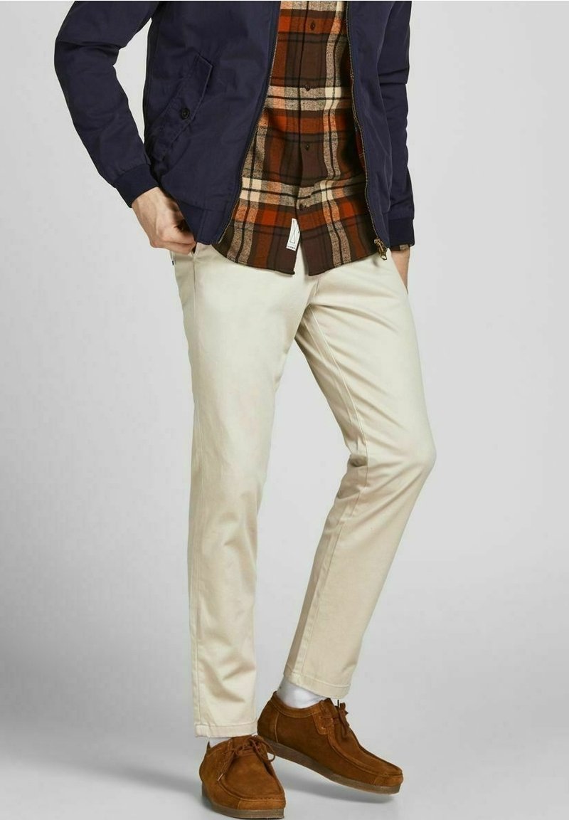 Beige chinos gecombineerd met een bruin en oranje geruit overhemd onder een marineblauwe bomberjack, aangevuld met bruine suède schoenen.