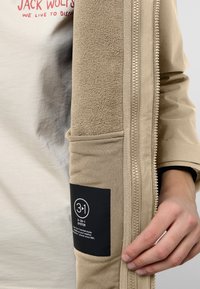 Giacca beige 3-in-1 con rivestimento in pile e chiusura con zip. Presenta una grande tasca e un'etichetta che indica il sistema multifunzionale.