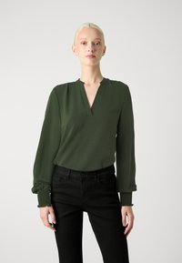 Zelená blouse s dlouhým rukávem, V-výstřihem a nařasenými manžetami, doplněná o černé kalhoty. Tkanina je měkká, což vytváří volný střih.