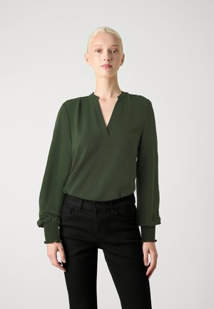 Grønn langermet bluse med V-hals og rynkede mansjetter, paret med svarte bukser. Stoffet er mykt og gir en avslappet passform.