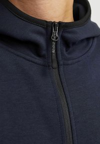 Marineblaue Zip-Hoodie mit hohem Kragen, ausgestattet mit einem schwarzen Reißverschluss und einer glatten, weichen Textur. Nahtdetails sind an der Vorderseite sichtbar.