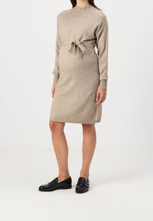 Pregnant woman wearing a beige knit sweater dress with a waist tie and black leather loafers, standing against a plain background.