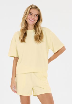 Smilende kvinde med langt blondt hår iført en løs lysegul kortærmet skjorte og matchende shorts mod en ensfarvet hvid baggrund.