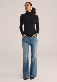 Femme aux cheveux bouclés portant des boucles d'oreilles blanches, un pull noir à col roulé, un jean bleu évasé et des chaussures noires à bout pointu, les mains dans les poches.