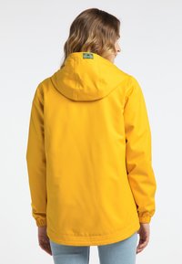 Gul vattentät jacka med huva, elastiska ärmslut och rak passform. Har en logotyp på baksidan. Slät textur, avslappnad design.