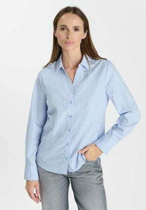 Vrouw met lang bruin haar die een lichtblauw gestreept overhemd met knopen en een grijze broek draagt, met één hand in haar zak.