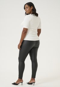 Frau in einem enganliegenden weißen Kurzarmoberteil und schwarzen glänzenden Leggings, kombiniert mit offenen schwarzen High Heels. Einfaches, elegantes Design.