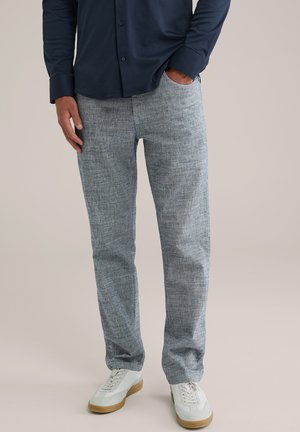 Pantalones de un azul claro, con textura y un corte holgado. Combinados con zapatillas blancas y grises. Camisa oscura de botones parcialmente visible.