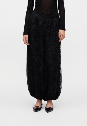 Femme portant un pantalon ample en dentelle noire avec des poignets élastiques et des ballerines noires pointues, debout devant un fond clair uni.