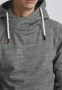 Sudadera gris con un acabado texturizado, que cuenta con un cuello alto, cordones blancos y detalles en tono beige en las puntas de los cordones.
