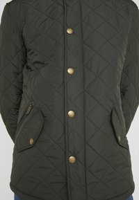 Quilted dark green jacket with a snap button closure, side pockets, and distinctive diamond pattern. Smooth texture with gold hardware accents.