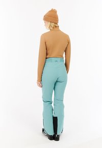 Bruine lange mouwen top, mintgroene skibroek met elastische tailleband, zwarte laarzenmanchetten, gezien vanuit de achterkant. Beaniemuts maakt de outfit compleet.
