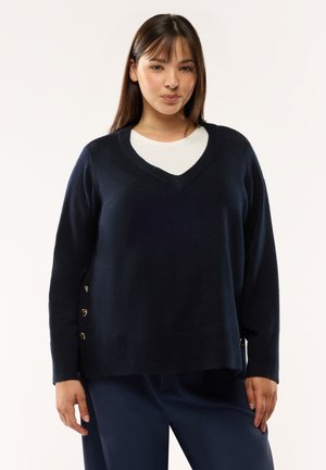 Giovane donna che indossa un maglione blu navy con scollo a V e bottoni laterali, sopra una maglietta bianca e pantaloni blu navy abbinati, in piedi davanti a uno sfondo semplice.