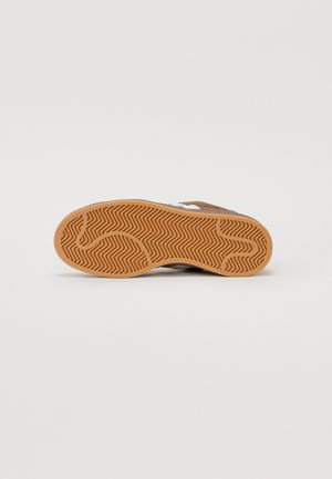 Baskets marron avec une semelle en caoutchouc gomme présentant un motif en chevrons, des accents blancs sur le côté, et un matériau supérieur en textile.